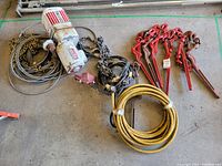 Complete lot overview showing the electric Coffing hoist with attached chains, eight red lever chain binders, and coiled yellow electrical cable laid on concrete floor.