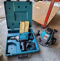 Makita electric drill in teal plastic carrying case with instruction manual and auxiliary handle; router positioned beside case