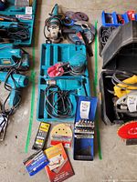 Photo showing Makita power tools including angle grinder, sander, and drill along with various accessories and drill bits on the ground.