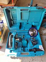 Open blue Makita plastic carrying case showing circular saw, charger with power cord, detachable handle, and blade fitting compartments.