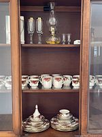 Cabinet shelf showing teacups, flutes, shot glasses and oil lamp
