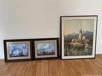 Three framed artworks displayed together: one large oil painting of a fishing boat and two smaller framed nautical prints leaning against the wall.