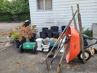 Overview of garden tools including wheelbarrow, various wooden-handled metal tools, black plastic pots, green lawn spreader, potted plants and decorative planters against house background.