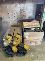 Two yellow Stanley Bostitch coil nailers and two boxes of metal corner plates stacked on the floor against a plywood wall