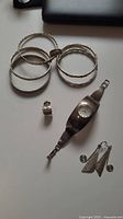 Four silver bangles with various textures and engravings, sterling silver watch, one silver and gold ring, and silver earrings spread on white surface.