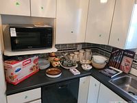 Wide shot of kitchen counter showing boxed Pyrex storage containers, ceramic mixing bowls, glass jars, and Panasonic microwave on shelf above.