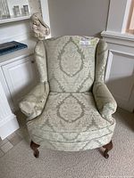 Front view of a wing back chair upholstered in light sage and beige damask fabric with wooden cabriole legs and armrest covers.