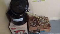 Photo showing assorted canning jars in cardboard boxes alongside a black enamel canner with lid on a box