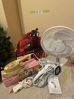 Red Dirt Devil handheld vacuum with the electrical cord wrapped around it, placed next to a boxed vintage Osrow Seamstress 11 steam iron, a white handheld electric scissor, a white handheld fabric steamer, and a small Holmes table fan on carpet.