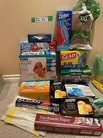 Full lot showing various household bags, Ziploc bags, Swiffer dusters, parchment paper, and aluminum foil on beige carpet with wall background.