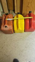 Photo of three gas jugs: orange, yellow, and red, arranged side by side with a measuring stick laid across them to show size.