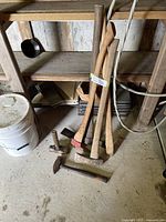 Overall view of the hand tools arranged on the ground near a wooden shelf, including axe, pick, and mini sledge hammers.