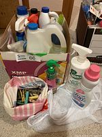 Photo showing various household cleaning products and boxed items, including detergent bottles, spray bottle, dispenser, and a plastic bag of clothes pins.