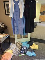 Blue dress with aquatic prints and white cardigan over it on hanger next to a black dress with sheer jacket, assorted folded shirts and tops below