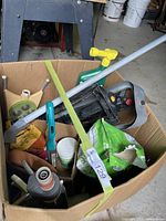 Box with assorted garden tools including hose nozzle, pruning clippers, green-handled weeder, various small tools, and bags of lawn seed and plant starter.