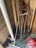 Photo showing a collection of long handled garden tools including rakes, pitchforks, a spade, and a water broom leaning against shed walls and pipe.