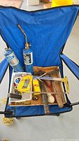 Lot contents displayed on a blue foldable chair including two blue butane gas bottles (one with torch attachment), a yellow retractable tape measure, hacksaw, coiled metal wire, metal file, and wooden handled tools.