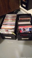 Two black handled storage boxes filled with assorted music CDs upright showing spines and cover art