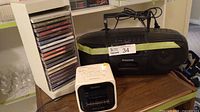 Photo showing Sony Dream Machine clock radio, Panasonic portable stereo CD player with power cord, and white CD rack with multiple music CDs.