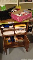 Wooden office cabinet with multiple compartments storing various paper office supplies and stapler on top