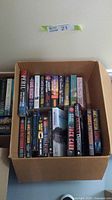 Box of assorted fictional adventure paperback books seen from above, titles visible include works by James Patterson, Jack Carr, and others.