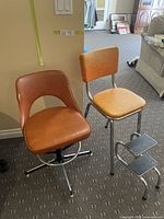Two vintage chairs side by side on carpeted floor: a chrome kitchen chair with orange vinyl upholstery and fold-out metal steps, next to a brown pleather swivel chair with chrome base.