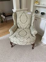 Front view of the wing back chair showing light sage damask patterned upholstery and wooden cabriole legs.
