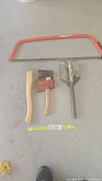 Four yard tools laid flat on concrete floor: a 30-inch Sandvik crosscut saw with orange frame, 12-inch hatchet with wooden handle and metal head with brown sheath, an extra wooden handle, and a multi-tool shovel with metal blade.