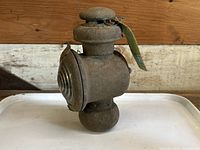 Front and side view of the 1914 Ford Model T kerosene parking lamp showing the metal body with aged patina and glass lens, resting on a white tray with a wooden background.
