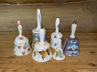 Photo of six porcelain and bone china bells with various floral and scenic designs arranged on wooden surface against rustic wood background.