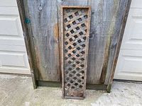 Front view of rectangular cast iron lattice window grate leaning against wooden fence showing rust and wear