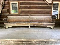 Extra-long antique church pew bench against a log wall on a porch, showing full length with wooden seat and backrest, cast iron ornate ends with scrollwork and floral motif.