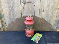Full view of vintage Coleman red lantern with glass globe and metal handle, placed on wooden surface with original Coleman lantern lighter in green packaging beside it.