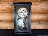 Front view of Gustav Becker wall clock, showing black wood case, clock face with black numerals, and lower glass panel.