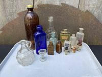 Full view of lot showing 11 apothecary bottles of various colors and shapes arranged on a white tray outdoors against a rusty background.