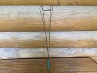 Front view of an antique wire carpet beater with a green-painted wooden handle resting against a wood log wall background.