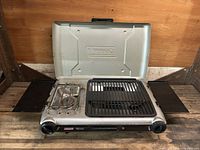 Coleman InstaStart Grill Stove open showing the single burner on the left and grill grate on the right with fold-out windblock panels.
