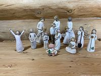 Group photo of all 11 Willow Tree figurines showing various poses and themes such as angels, mothers, children, sunflowers, and pets on a wooden surface with log cabin background.