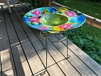 Glass birdbath with colorful embossed hummingbirds, butterflies, and flowers on painted metal stand outdoors.