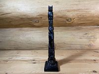 Front view of a tall, narrow, hand-carved wooden totem pole sculpture with distinct Indigenous-style figures and detailed patterns, standing on a square base against a log background.