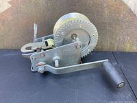 Side view of heavy-duty hand crank winch with large gear and crank handle, showing metal construction and attached yellow strap.