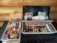 Two large plastic totes filled with assorted holiday and birthday wrapping supplies including gift bags, tissue paper, rolls of wrapping paper, ribbons, and gift tags in various patterns and colors.