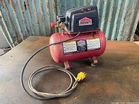 Jobmate 3-gallon air compressor placed on a table against a corrugated metal background. Shows full unit with red tank and black motor housing, power cord extended in front.