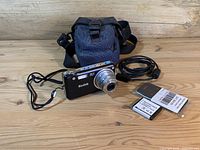 Kodak EasyShare V1253 camera with lens extended, showing included accessories of camera case, USB cable, two batteries, and charger on wooden surface.