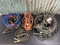 Large assortment of industrial cables including welding leads, booster cables, and hydraulic/pneumatic hoses laid out on a black surface against a corrugated metal background.