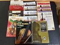 Overview photo showing all 14 violin and fiddle music books arranged on a surface. Titles visible include Favorite Celtic Melodies for Fiddle, Mel Bay’s Old Time Fiddle Solos, Medieval Music for Violin, and multiple Violin Series instructional books plus genre-repertoire books.