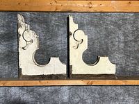 Front view of two antique wooden architectural corbels with worn chippy white paint showing ornate scroll design and natural patina, placed on wooden framing against gray backdrop.