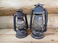 Both vintage kerosene lanterns side by side on wooden surface, showing size difference and overall condition