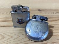 Two vintage metal pocket lighters on a wooden surface, one rectangular with a heraldic shield emblem, the other rounded with engraved stripes and monogram.