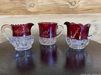 Three ruby flash pressed glass pieces including two creamers and one handled cup displayed together on a wooden surface.